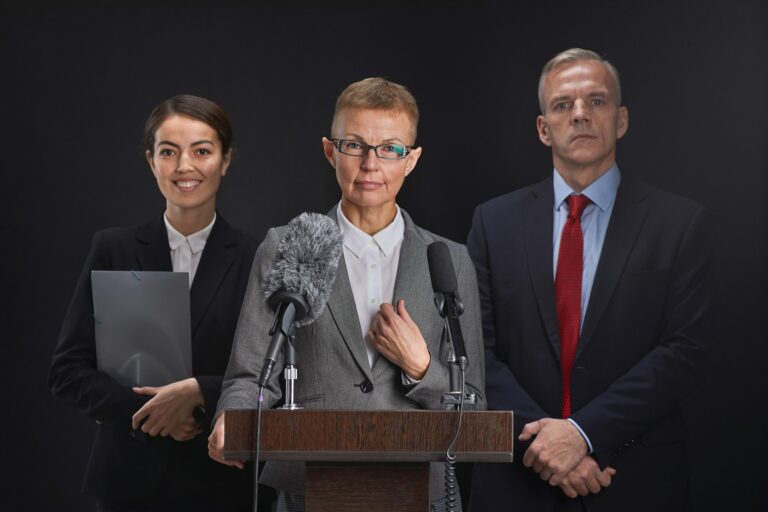 Portrait à la taille d'une femme mûre prononçant un discours debout sur le podium avec deux assistants en arrière-plan