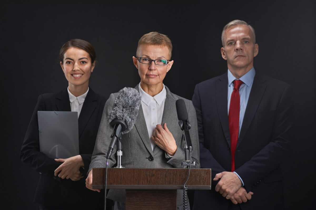 Portrait à la taille d'une femme mûre prononçant un discours debout sur le podium avec deux assistants en arrière-plan