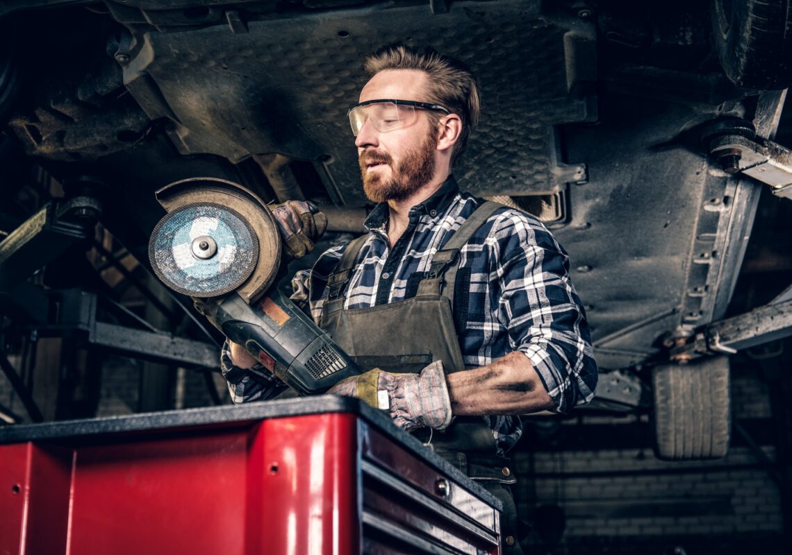 Un mécanicien barbu portant des lunettes de protection tient une meuleuse d'angle près de la voiture dans un atelier.