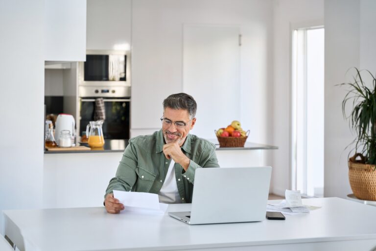 - 22 Heureux homme d'âge moyen entrepreneur portant des lunettes regardant une facture papier vérifiant une facture financière ou un document fiscal effectuant des paiements en ligne en travaillant sur un ordinateur portable à la table de la cuisine à la maison.