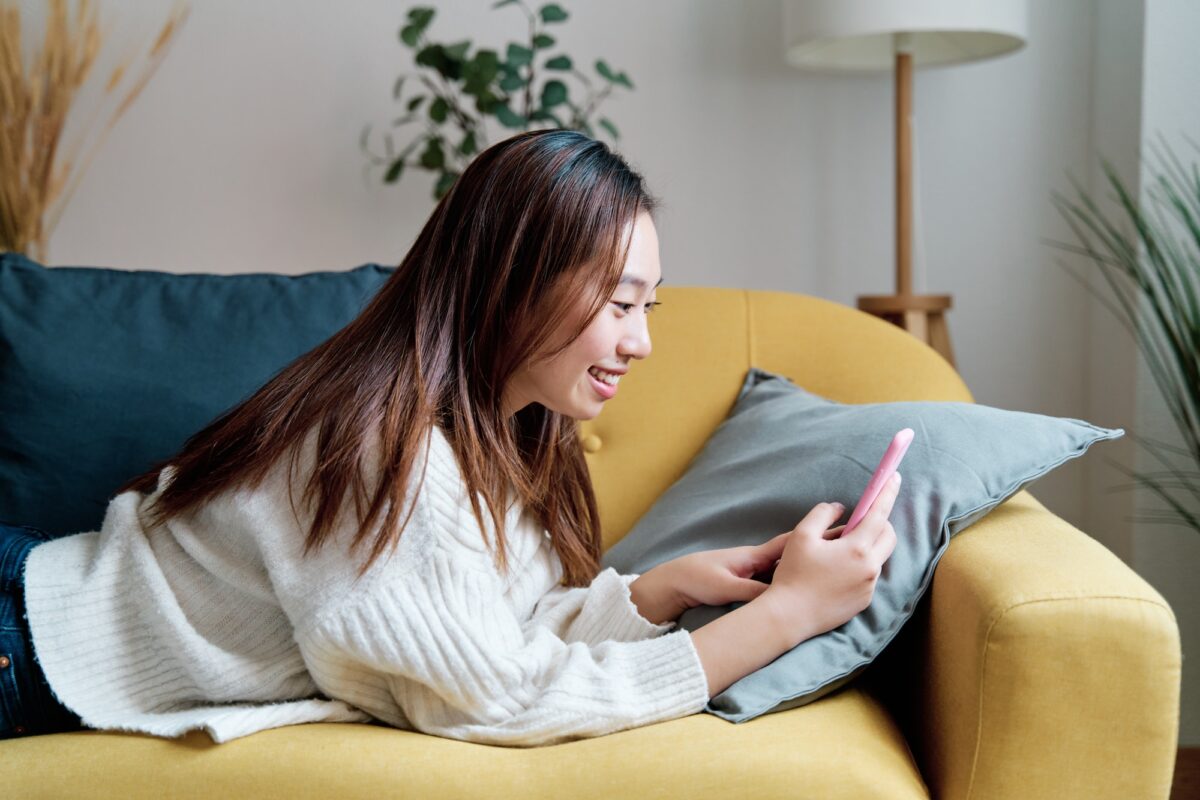 Jeune femme positive dans des vêtements décontractés souriant et parcourant son téléphone portable tout en se reposant sur un canapé dans un salon confortable à la maison