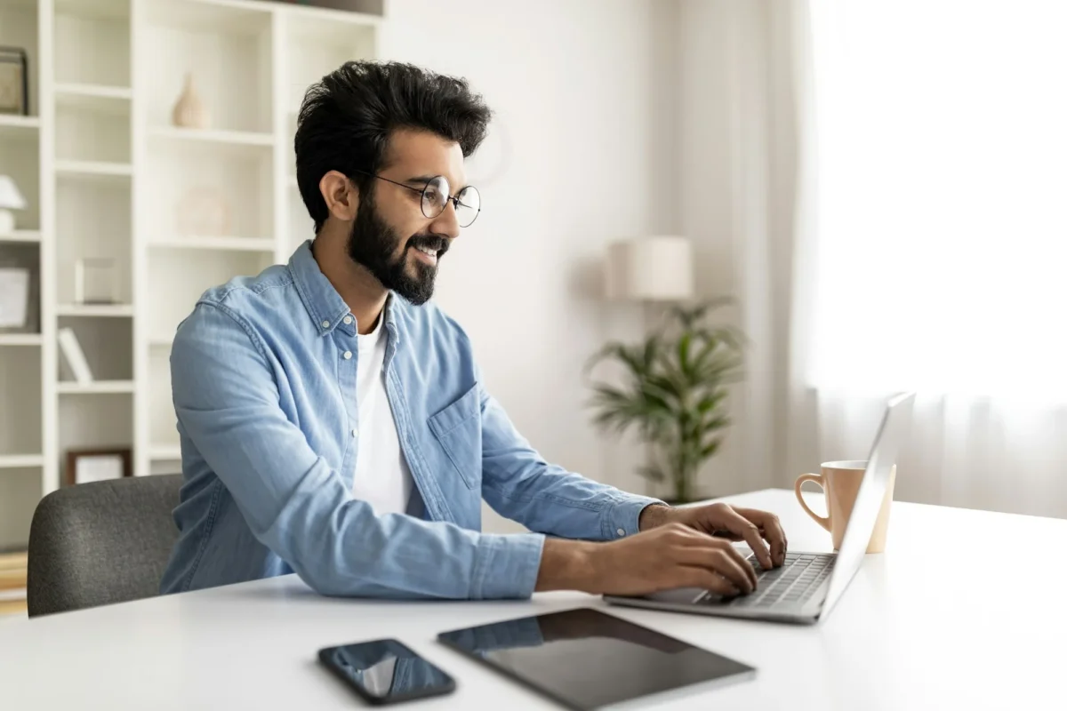- 1 Indépendant indien souriant travaillant avec un ordinateur portable au bureau à domicile, bel homme oriental assis au bureau avec un ordinateur, une tablette numérique et un smartphone, utilisant des gadgets modernes pour le travail, espace de copie